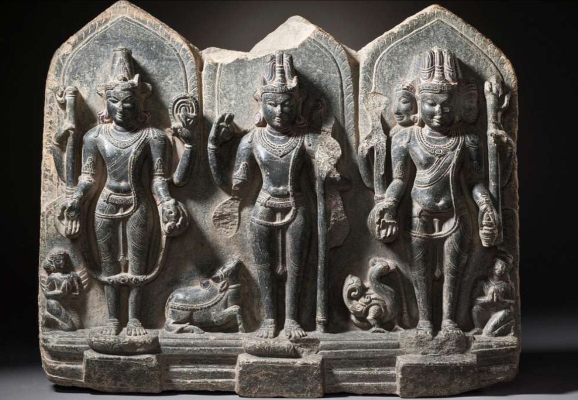 Stone relief of the Trimūrti — Brahmā, Viṣṇu and Śiva standing side by side in a triple-arched shrine, with their vāhanas at their feet.
