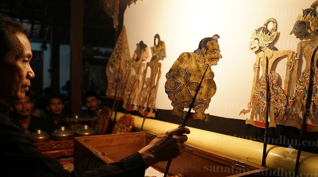 Close study of the Javanese Punokawan puppet Semar — short, heavy-bodied, plain-featured, carved in leather and painted in muted gold and black — held up against the glowing screen while the refined Kṣatriya puppets stand behind him.